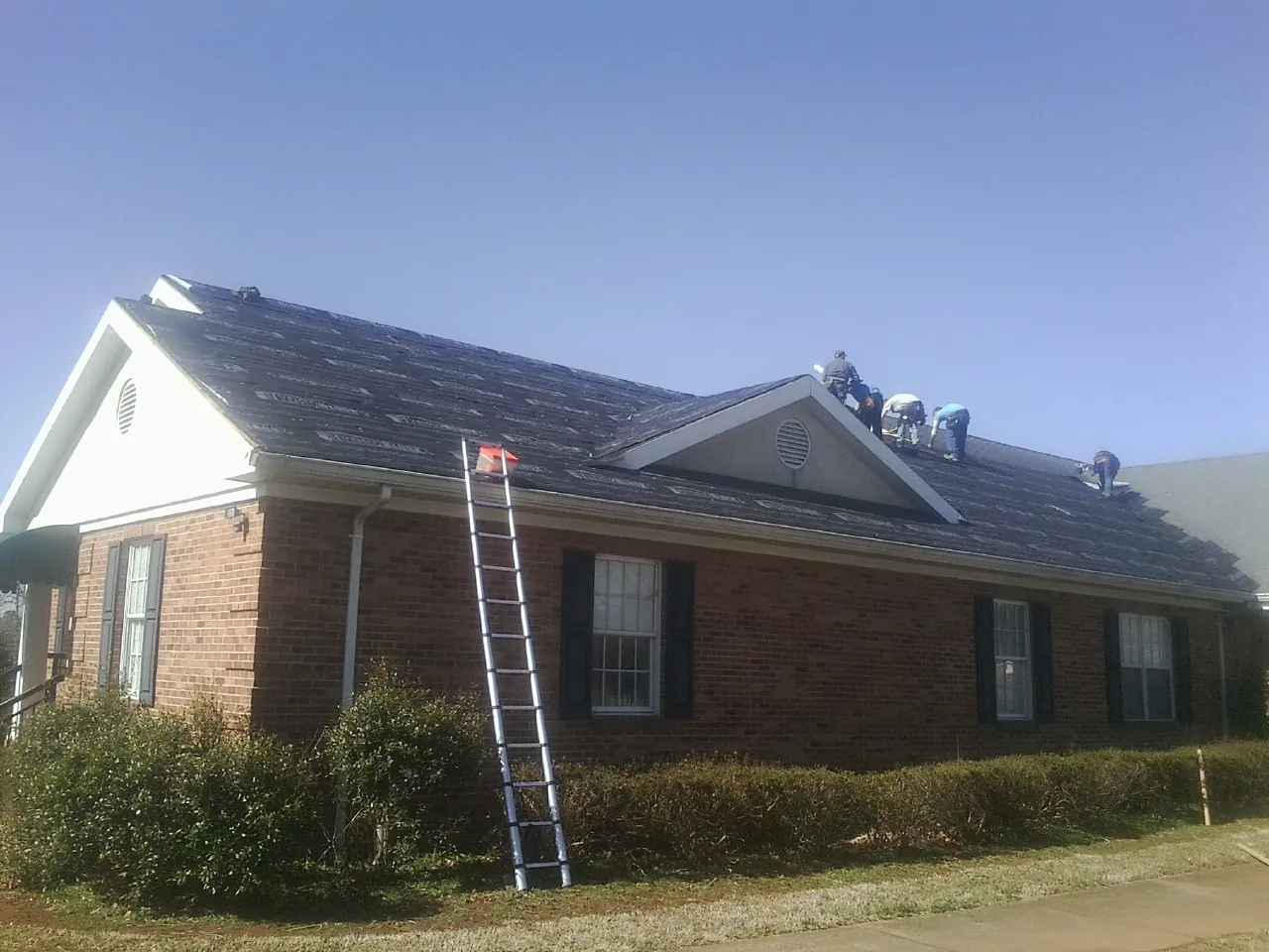 Crew tearing shingles off a brick ranch roof with a ladder set — Emergency Roof Repair work by our crew in Gilford
