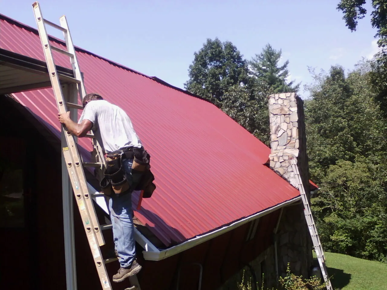 Roofer on a ladder beside a red metal roof and stone chimney — Soffit & Fascia Repair by Roofing Brothers in Gilford